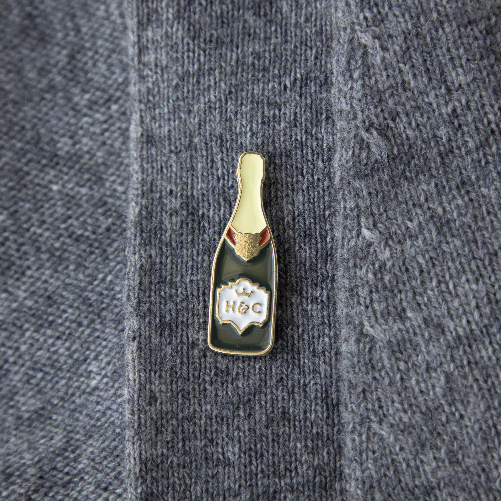 A gilded story envelops a bottle of Hester &amp; Cook Champagne Enamel Pin on a marble table.