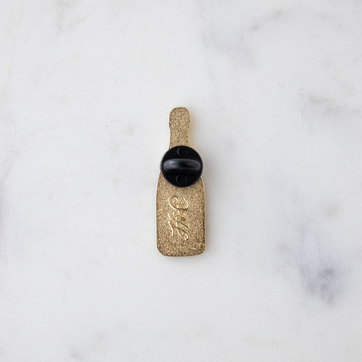 A gilded story envelops a bottle of Hester &amp; Cook Champagne Enamel Pin on a marble table.