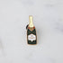 A gilded story envelops a bottle of Hester & Cook Champagne Enamel Pin on a marble table.