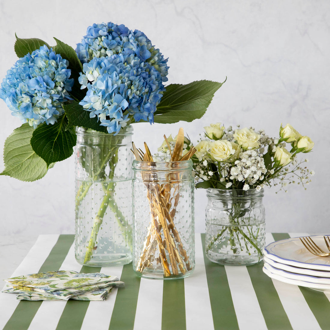Three glass Clear Hobnail Jars sit on the Dark Green Classic Stripe runner, one with blue hydrangeas, one with white flowers, and one with gold utensils. Plates and floral napkins complete the arrangement on the side.