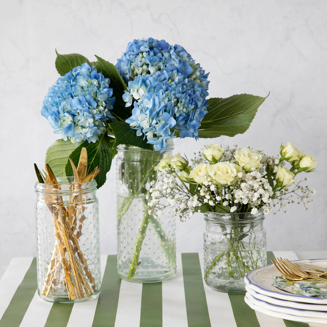 Three glass Clear Hobnail Jars sit on the Dark Green Classic Stripe runner, one with blue hydrangeas, one with white flowers, and one with gold utensils. Plates and floral napkins complete the arrangement on the side.