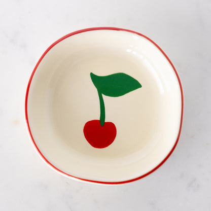 A white ceramic pinch bowl with a red rim features a painted bright red cherry with a green leaf.
