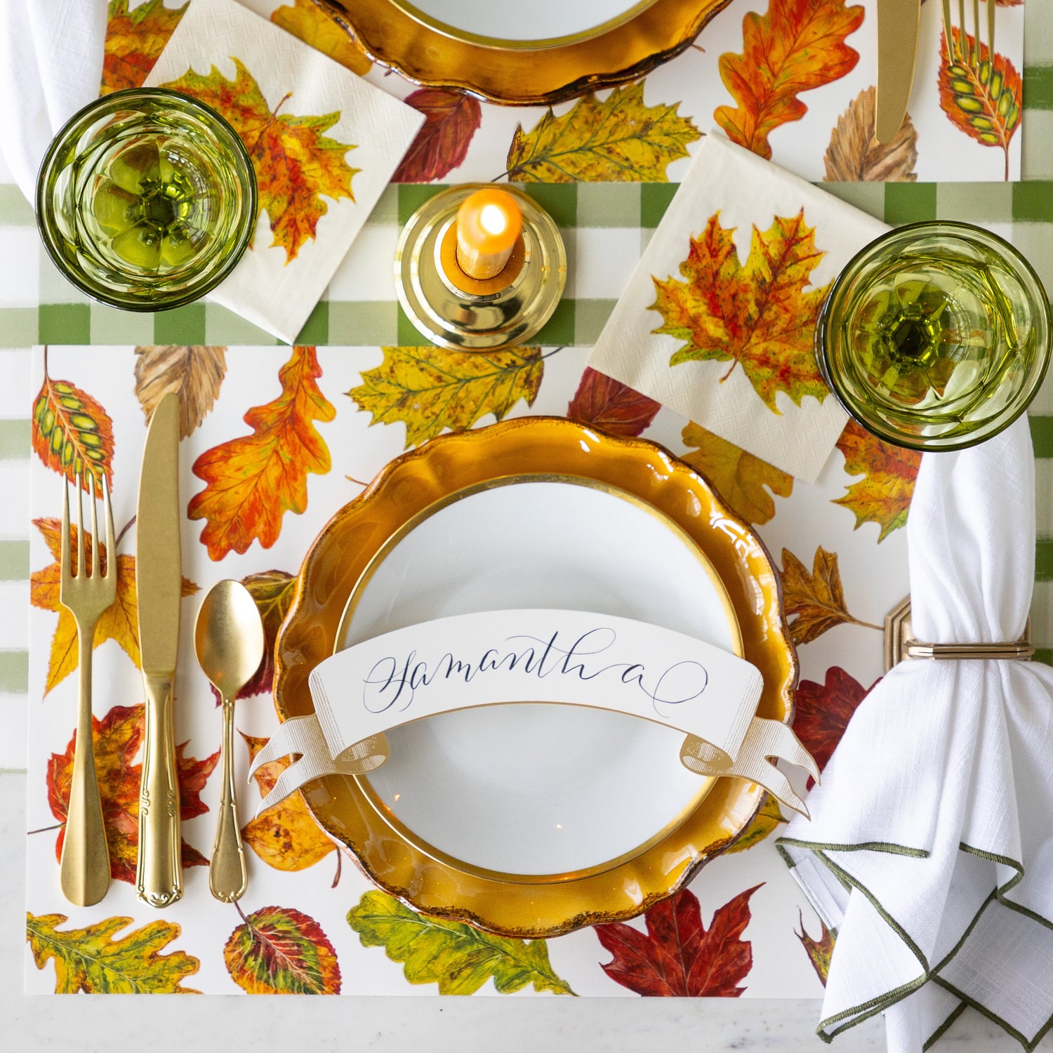 A place setting featuring the Fall Foliage Placemat under Mepra gold flatware, dinnerware with the Classic Gold Banner Table Accent reading &quot;Samantha&quot;, atop, a  napkin in a holder, the Fall Foliage Cocktail Napkin under a glass and Moss Green Painted Check Runner underneath.