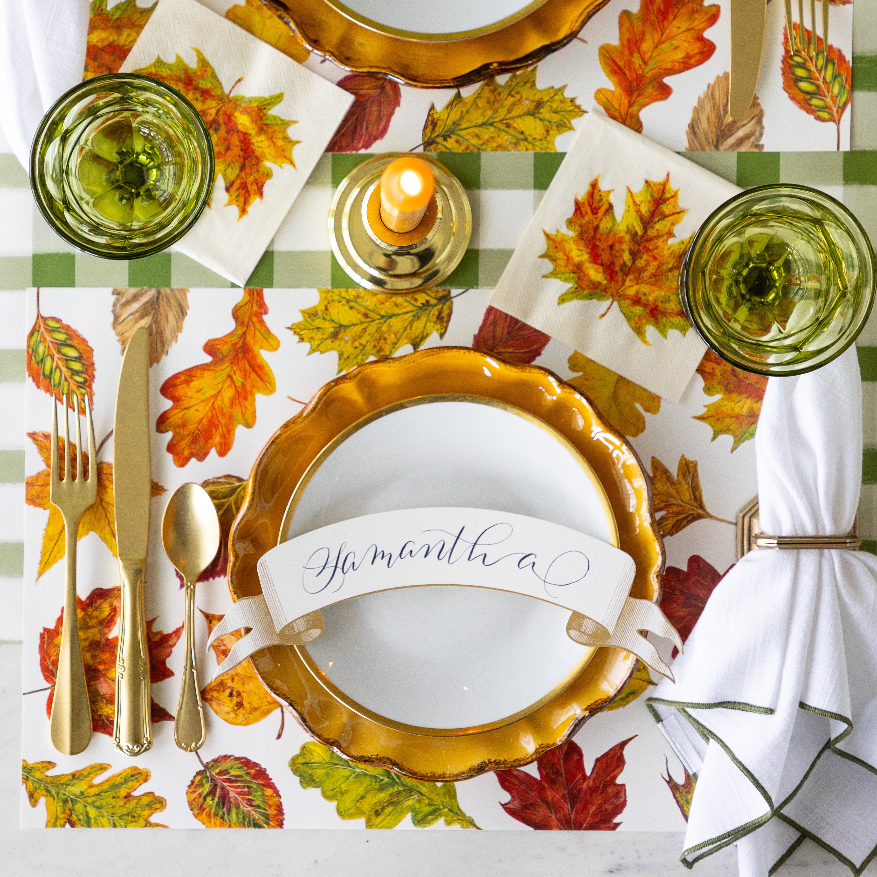 A place setting featuring the Fall Foliage Placemat under Mepra gold flatware, dinnerware with the Classic Gold Banner Table Accent reading &quot;Samantha&quot;, atop, a  napkin in a holder, the Fall Foliage Cocktail Napkin under a glass and Moss Green Painted Check Runner underneath.