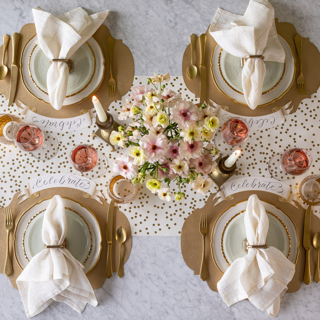 An elegant table setting for four with a floral centerpiece and lit candles, featuring the Die-cut Gold French Frame Placemats with gold rimmed dinnerware, gold flatware, glasses of wine, Gold Banner Table Accents reading &quot;Celebrate&quot; and Gold Confetti Runner underneath.