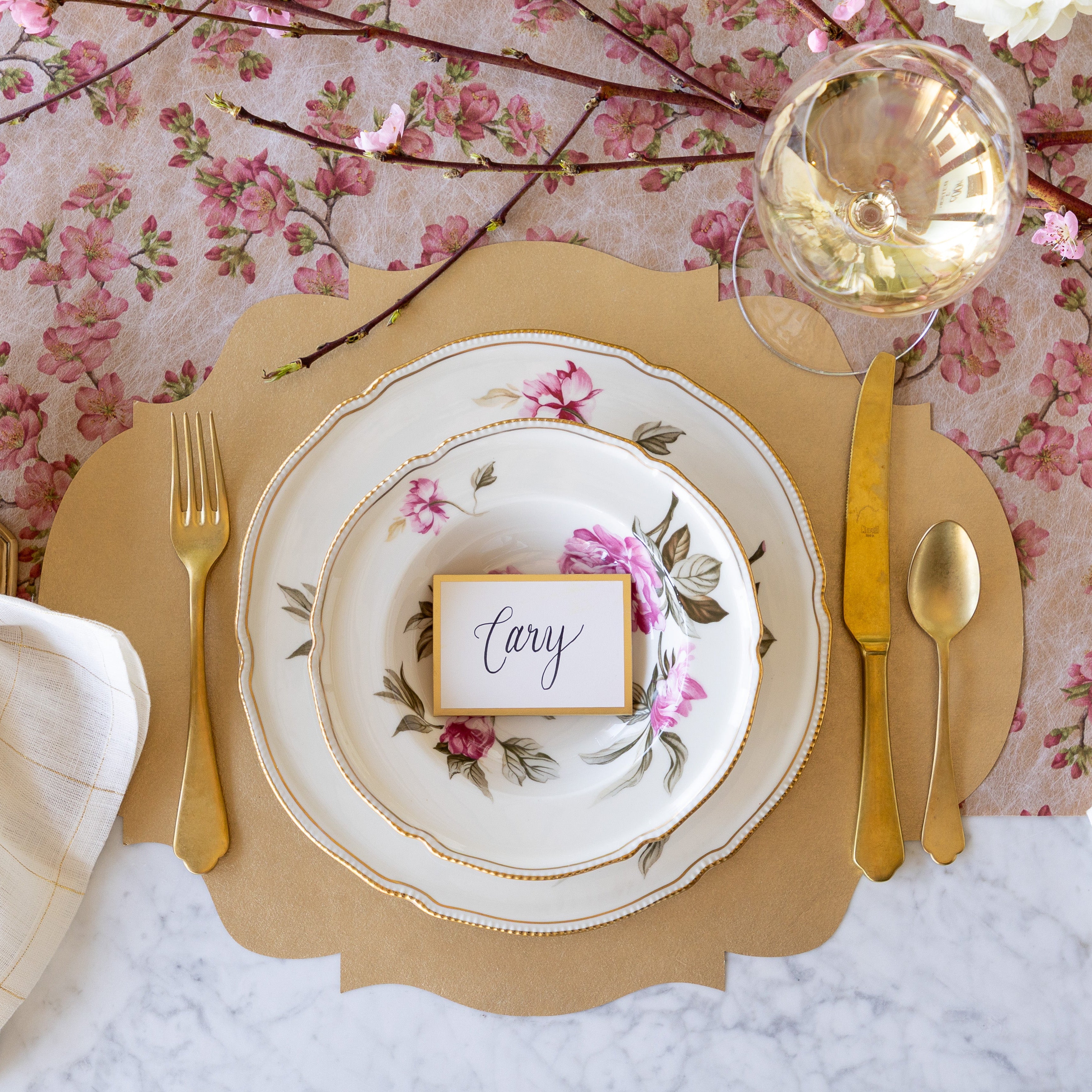An elegant place setting featuring the Die-cut Gold French Frame Placemat, vintage dinnerware with the Gold Frame Place Card reading &quot;Cary&quot; atop, gold flatware, glass of wine, and Cherry Blossom Runner underneath.