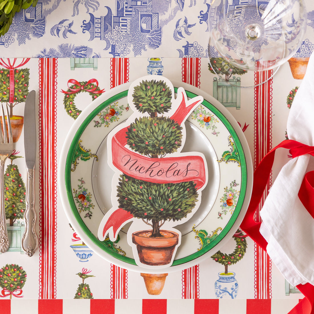An elegant place setting featuring the Holiday Topiary Placemat and Holiday Topiary Table Accent with &quot;Nicholas&quot; written in script on the ribbon of the table accent.