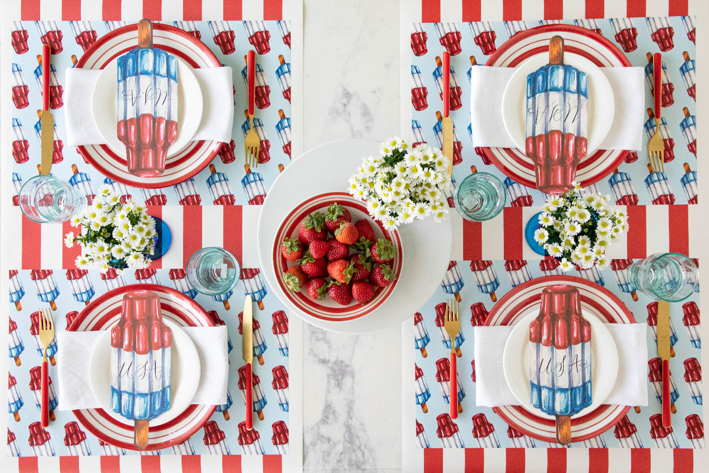 A patriotic table setting with flowers and strawberries in the center, featuring the Rocket Pop Placemat, red and white plates, Rocket Pop Table Accent reading "U.S.A" and red and gold flatware on the Red Classic Stripe Runner.