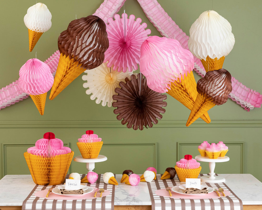 Paper honeycomb in the shapes of ice cream cones and wheels hand above a table that has cupcake shaped honeycomb and small ice cream cone shaped honeycomb.