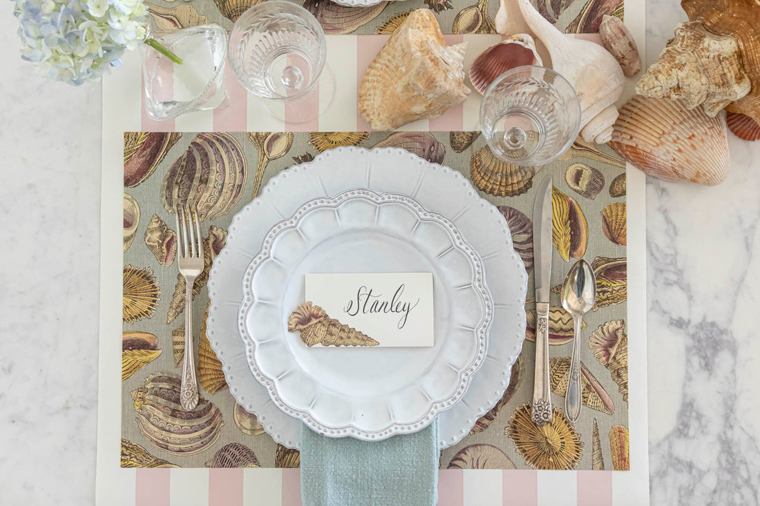 Place setting with a placemat that has shells printed all over it with white dinner plate and salad plate on top.  The table is decorated with pink & white striped paper table runner and shells scattered on it. 