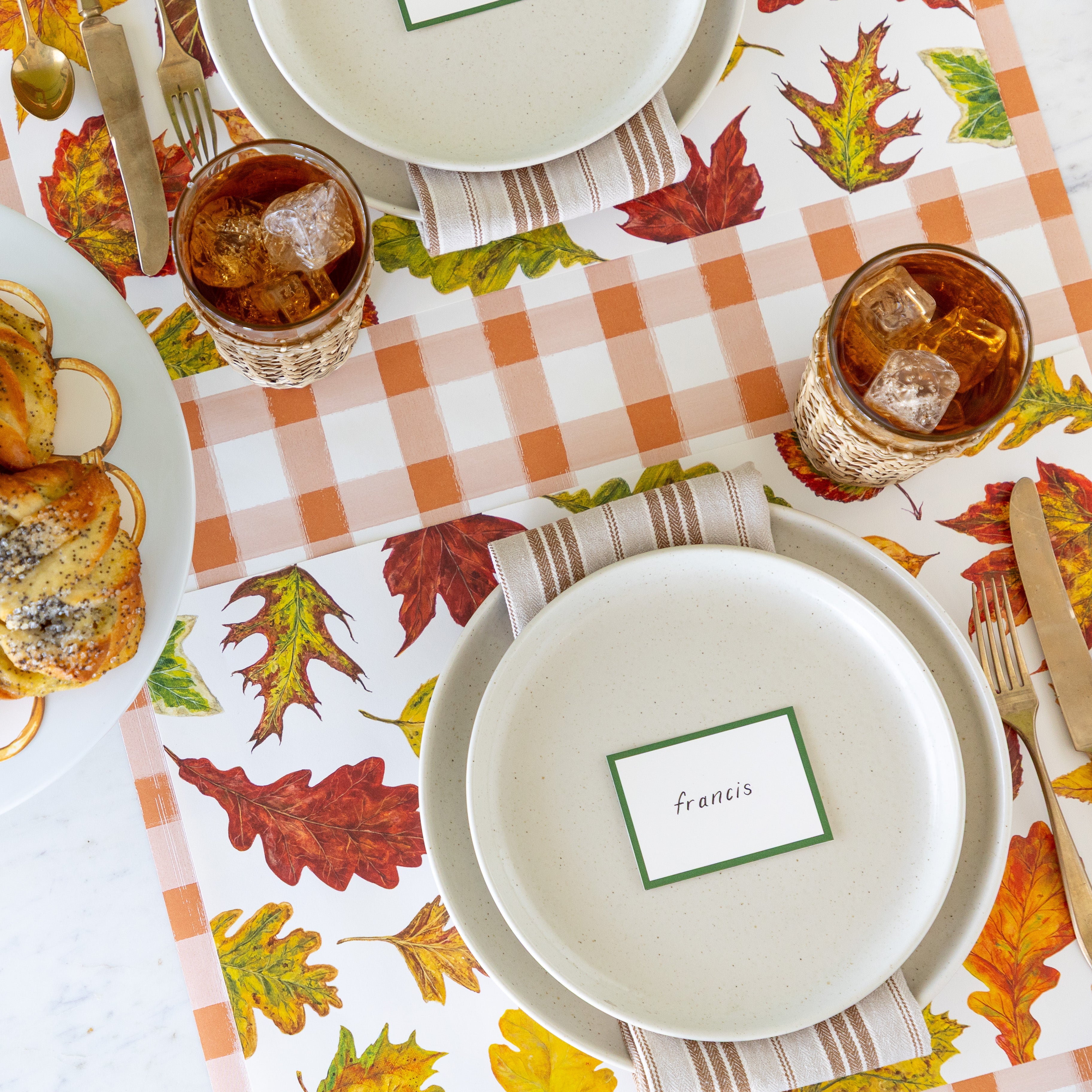 An elegant place setting featuring the Fall Foliage Placemat with a brown striped napkin in between two dinner plates with the Dark Green Frame Place Card reading &quot;francis&quot;, on top, a glass of iced tea and Orange Painted Check Runner underneath.