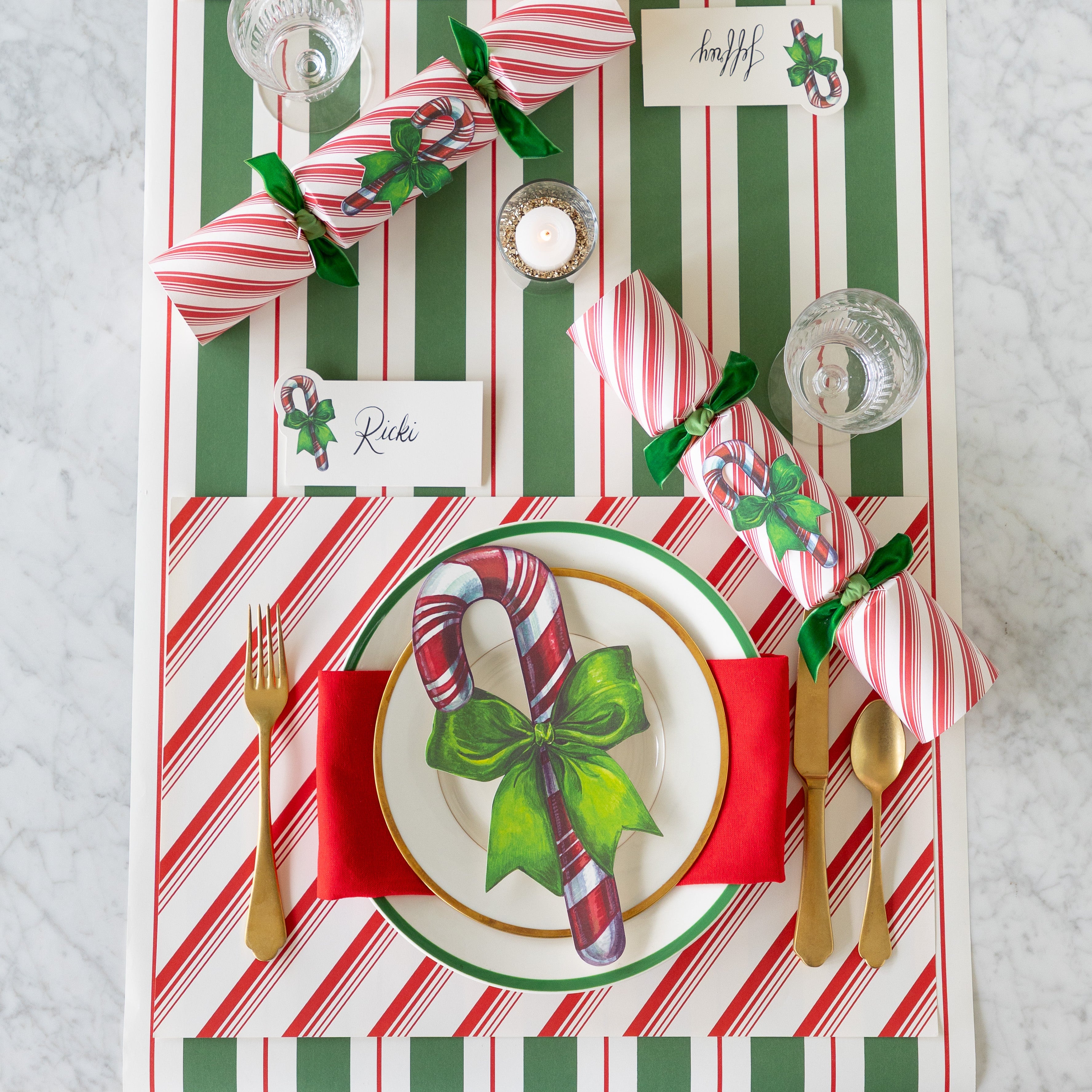 A place setting featuring the Peppermint Stripe Placemat, gold flatware, gold and green trimmed plates with the Candy Cane Table Accent atop, Candy Candy Place Card, Candy Cane Crackers and Green &amp; Red Awning Stripe Runner underneath.
