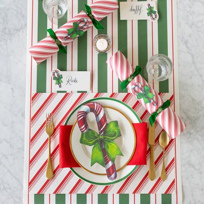 A place setting featuring the Peppermint Stripe Placemat, gold flatware, gold and green trimmed plates with the Candy Cane Table Accent atop, Candy Candy Place Card, Candy Cane Crackers and Green &amp; Red Awning Stripe Runner underneath.