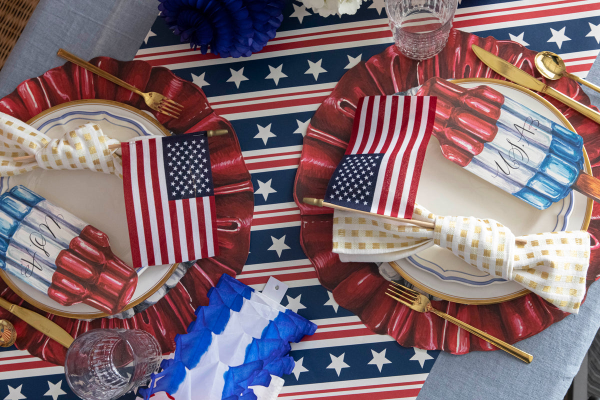 A Patriotic table setting featuring the Die-cut Star Spangled Placemat, Rocket Pop Table Accents, American flags tucked in gold-and-white napkins, and gold flatware on the Stars & Stripes Runner.