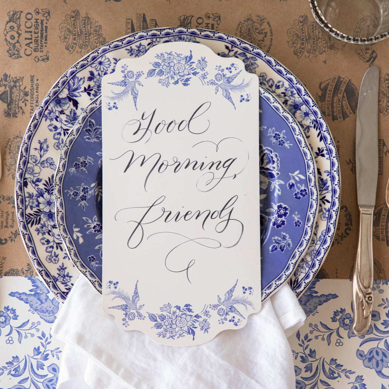 Blue Asiatic Pheasants Table Card with &quot;Good Morning Friends&quot; written on it, resting in the center of dinner plates, on a place setting for one.