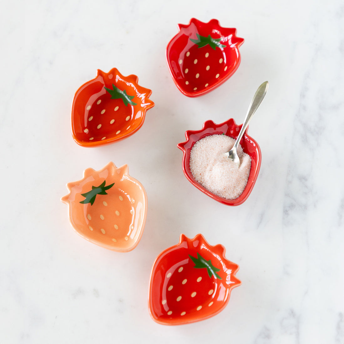 Five theme-shaped bowls resembling strawberries—four red and one peach-colored—are arranged on a white surface; one red dish holds a spoon and sugar.