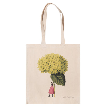 The Annabelle Heavyweight Canvas Tote Bag is light tan featuring a stylized illustration of a woman holding a gigantic green hydrangea bloom by the stem.