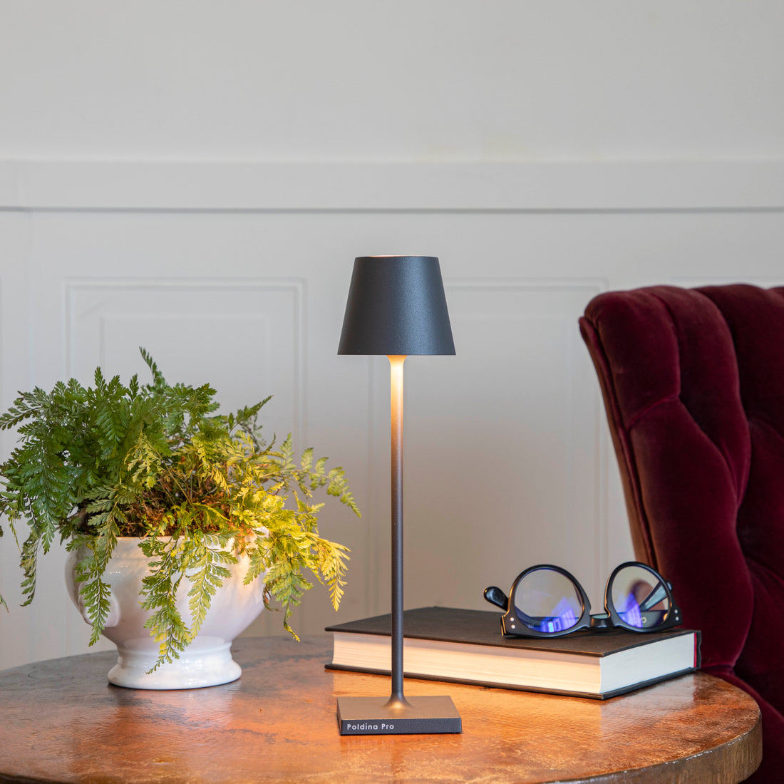 A Zafferano Dark Grey Micro Cordless Lamp on a table next to a book and a plant.