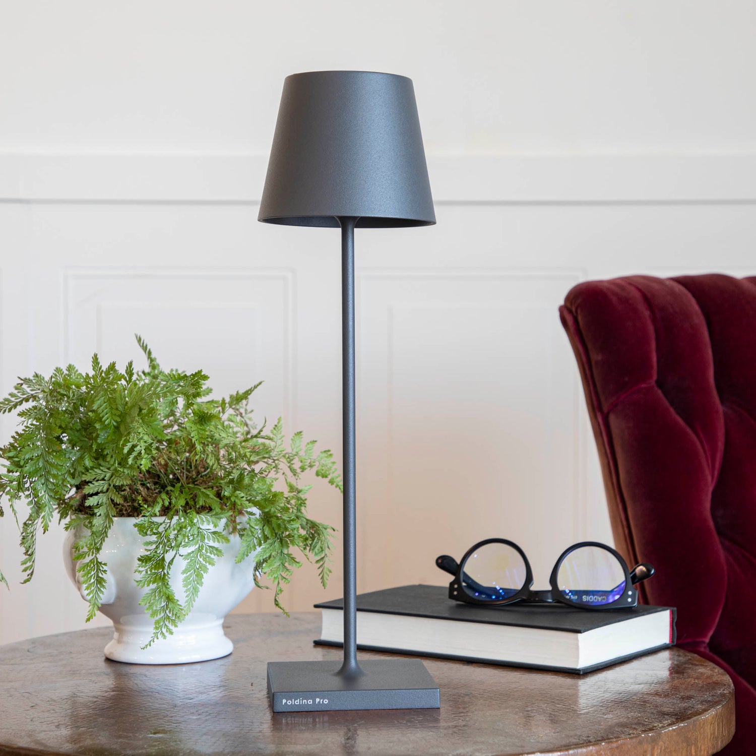 A touch-dimming control Zafferano Dark Grey Cordless Lamp on a table next to a book and a plant.