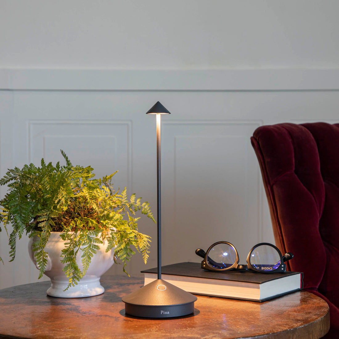 A Dark Grey LED Cordless Lamp by Zafferano on a table next to a book and a plant.