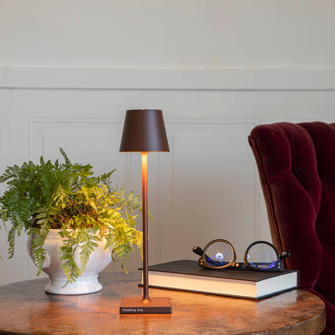 A cozy reading nook with a Zafferano Rust Micro Cordless Lamp, a green potted fern, and a pair of round glasses resting on an open book.