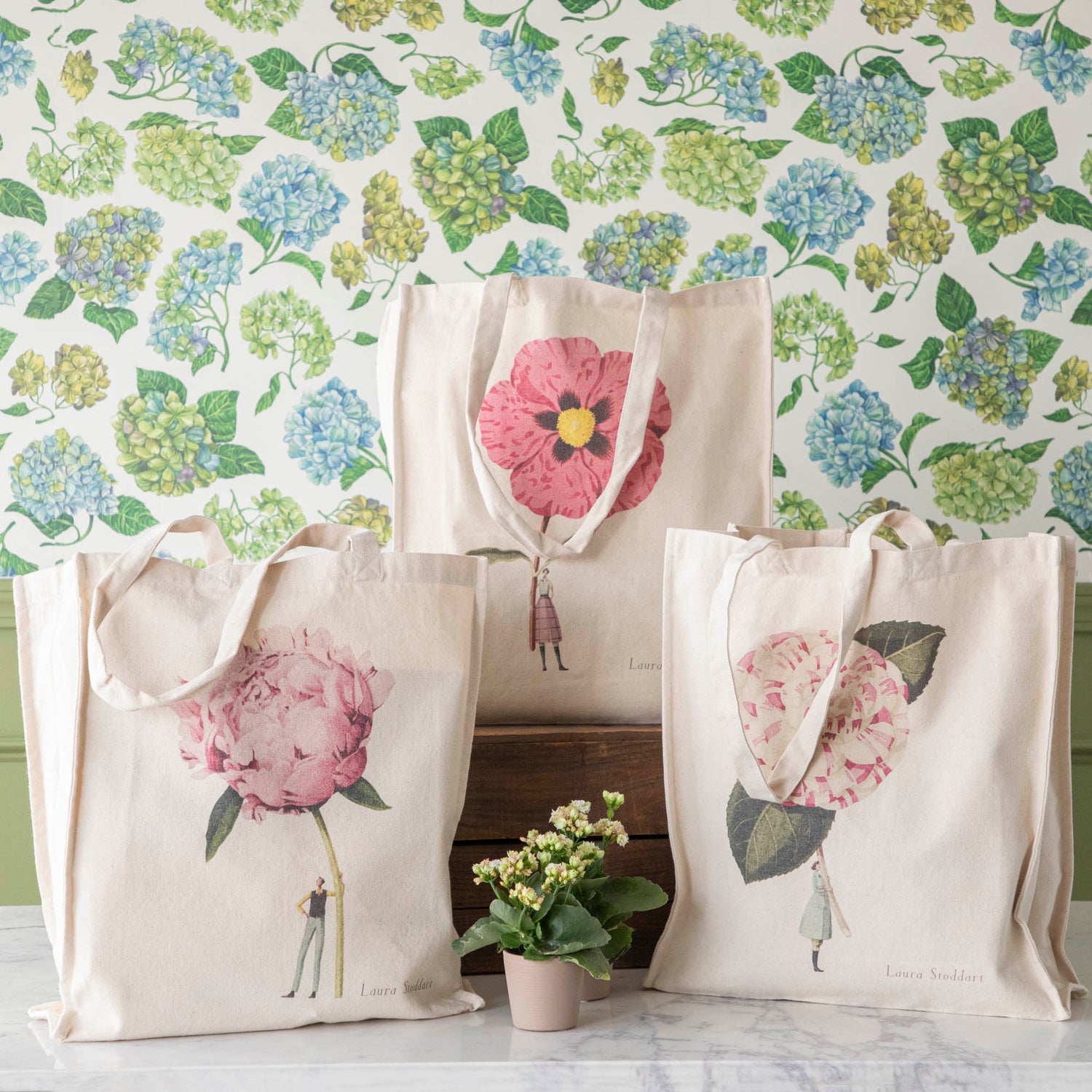 Three of the Heavyweight Canvas Tote Bags featuring pink floral artwork by Laura Stoddart arranged alongside potted plants against a hydrangea wallpaper background.