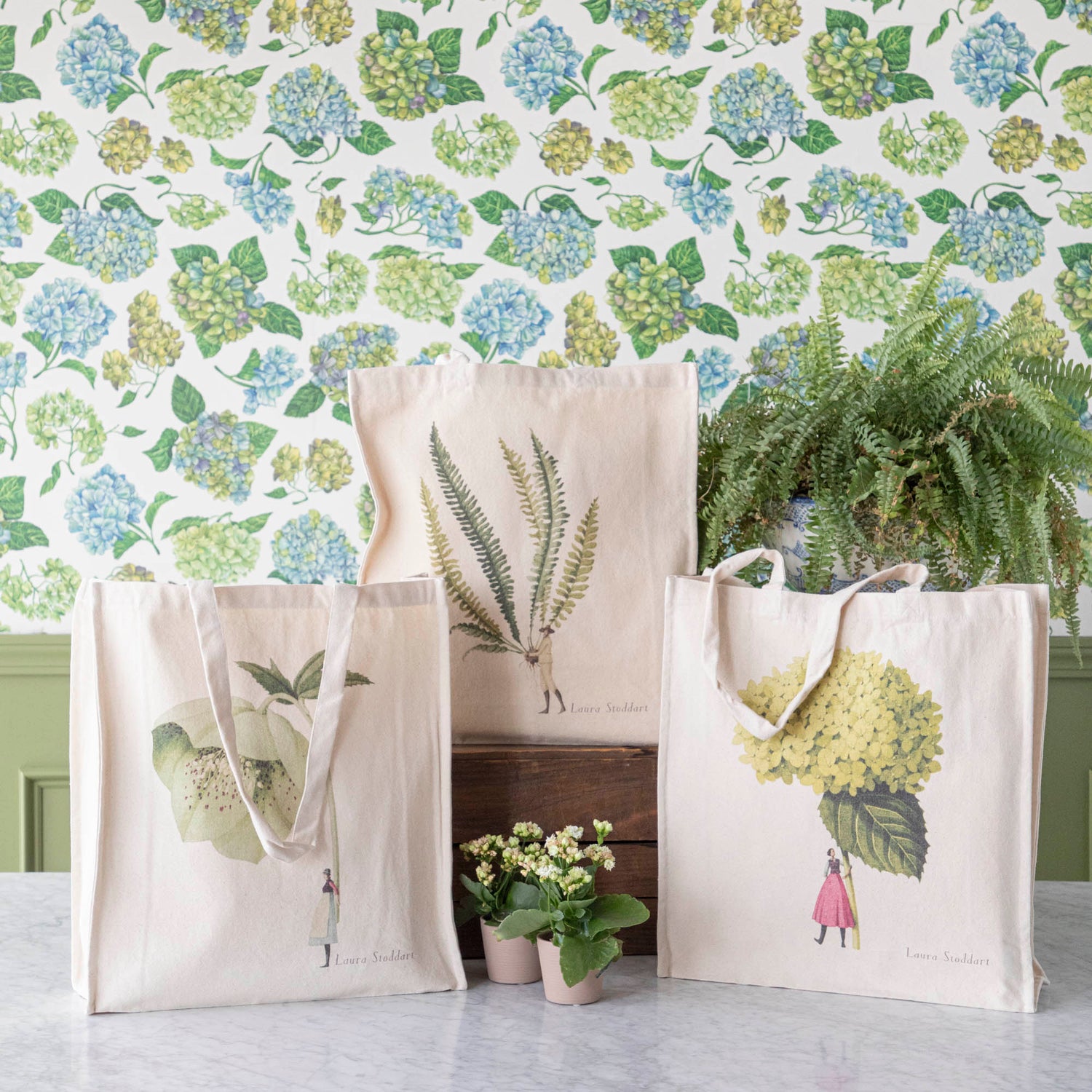 Three of the Heavyweight Canvas Tote Bags featuring green botanical artwork by Laura Stoddart arranged alongside potted plants against a hydrangea wallpaper background.