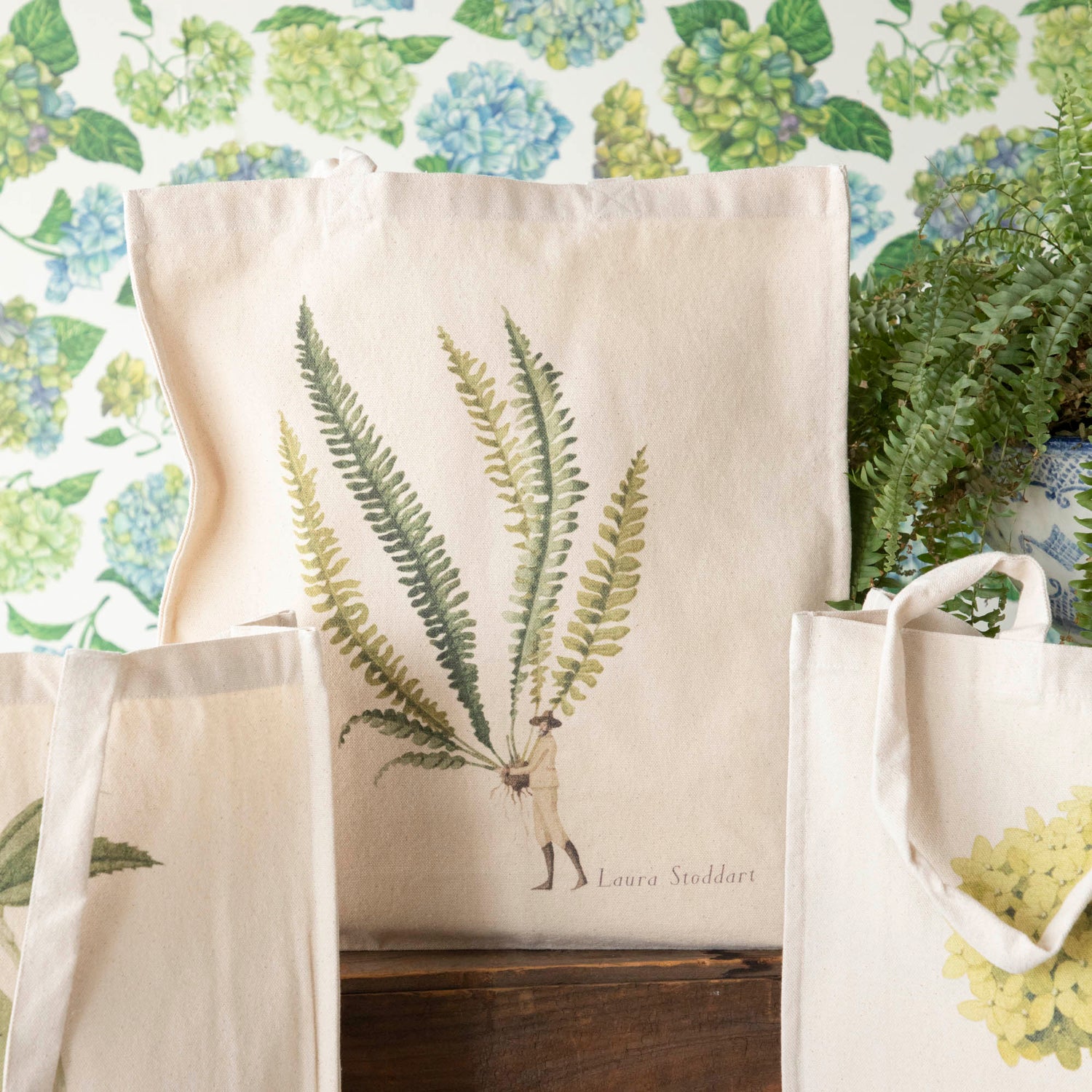 Close-up of the Fern 3 Heavyweight Canvas Tote Bag standing upright in a display, showing the beautiful illustration of a man holding a gigantic fern frond by the stem.