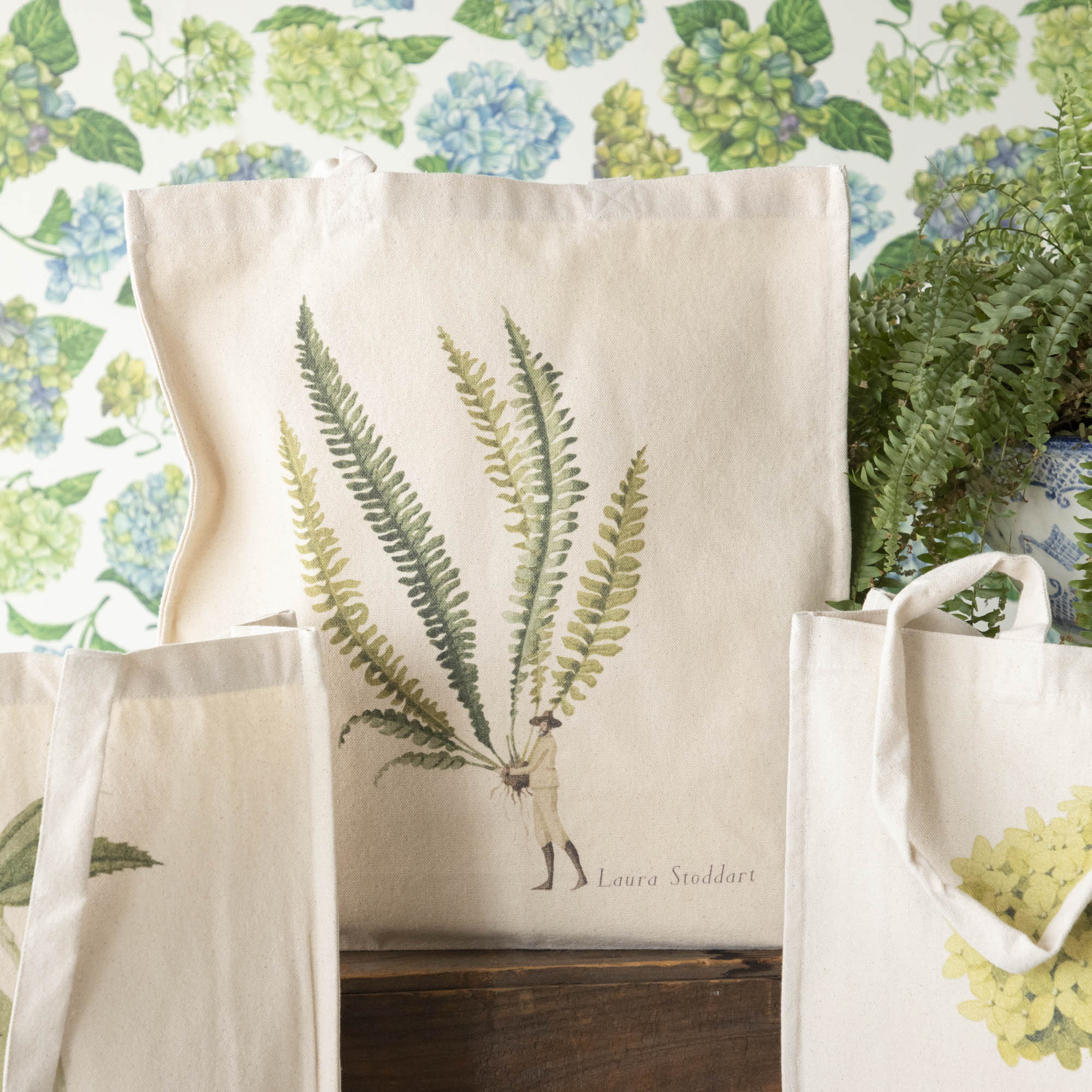 Close-up of the Fern 3 Heavyweight Canvas Tote Bag standing upright in a display, showing the beautiful illustration of a man holding a gigantic fern frond by the stem.