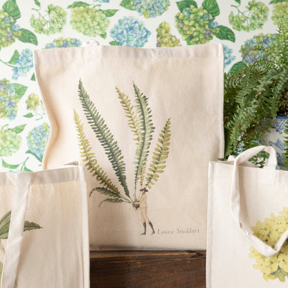 Close-up of the Fern 3 Heavyweight Canvas Tote Bag standing upright in a display, showing the beautiful illustration of a man holding a gigantic fern frond by the stem.