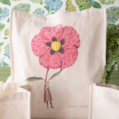Close-up of the Cistus Heavyweight Canvas Tote Bag standing upright in a display, showing the beautiful illustration of a woman holding a gigantic pink flower.
