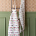 Two Laura Stoddart Aprons by Hester & Cook hanging on a wooden ladder against a wall with patterned wallpaper above wainscoting.