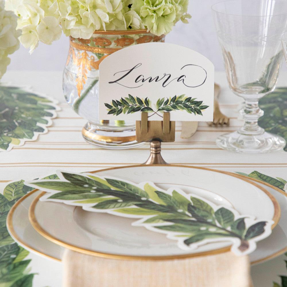 An elegant place setting featuring a Laurel Place Card labeled &quot;Laura&quot; in a brass place card holder behind the plate.