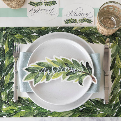 An elegant place setting featuring a Laurel Table Accent with &quot;Welcome&quot; written on it in white ink resting on the plate, from above.