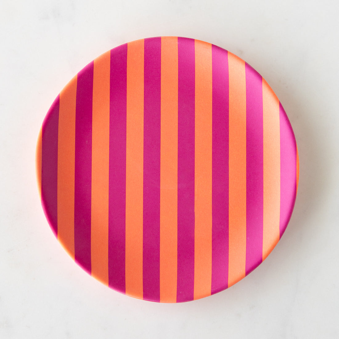 A round Stripe Melamine Plate with alternating vertical orange and magenta stripes on a marble table.