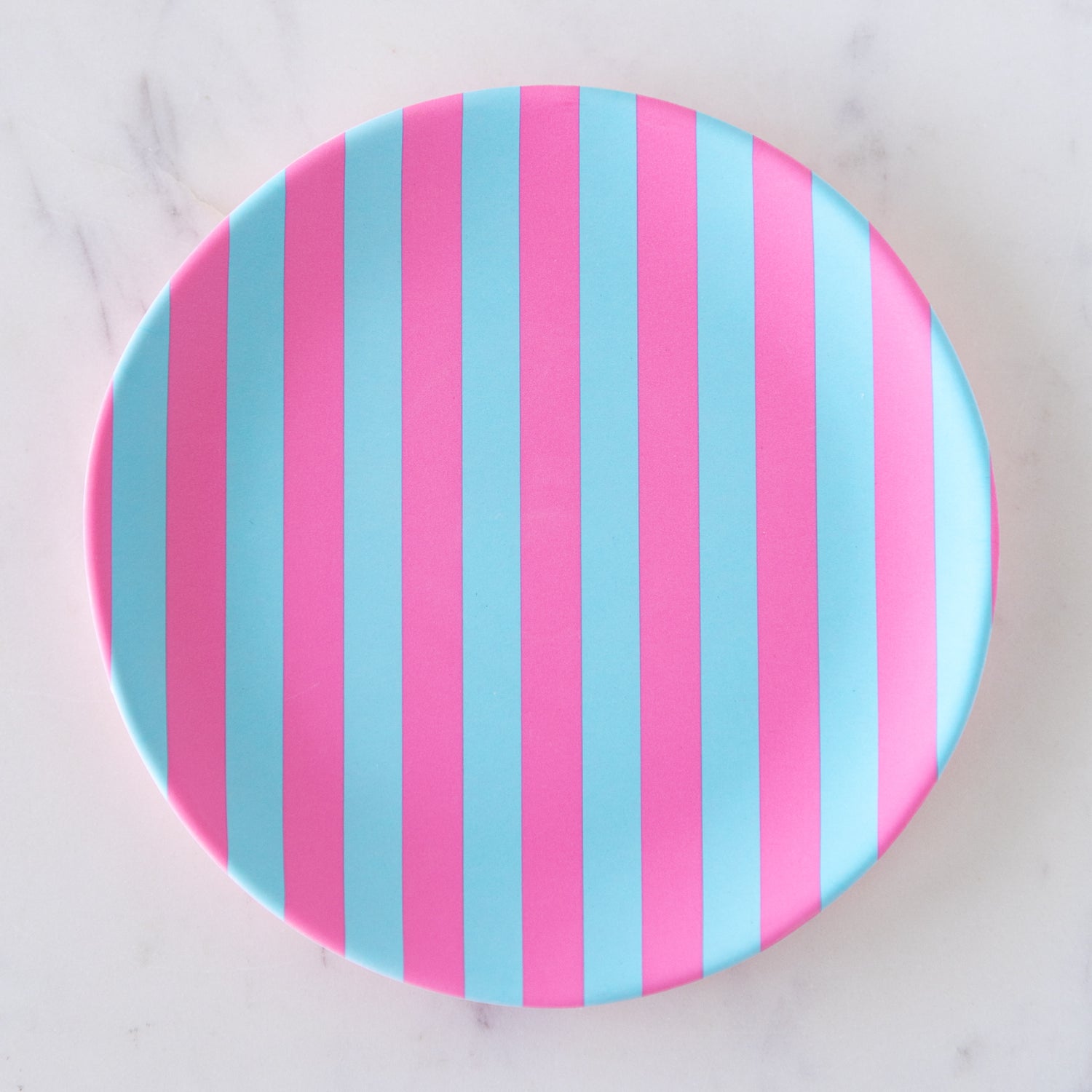 A round Stripe Melamine Plate with alternating vertical pink and light blue stripes on a marble table.