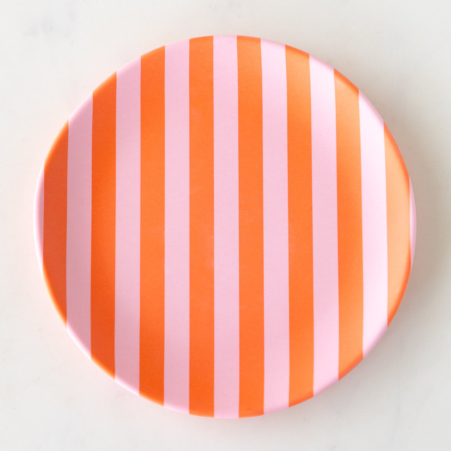 A round Stripe Melamine Plate with alternating vertical pink and orange stripes on a marble table.