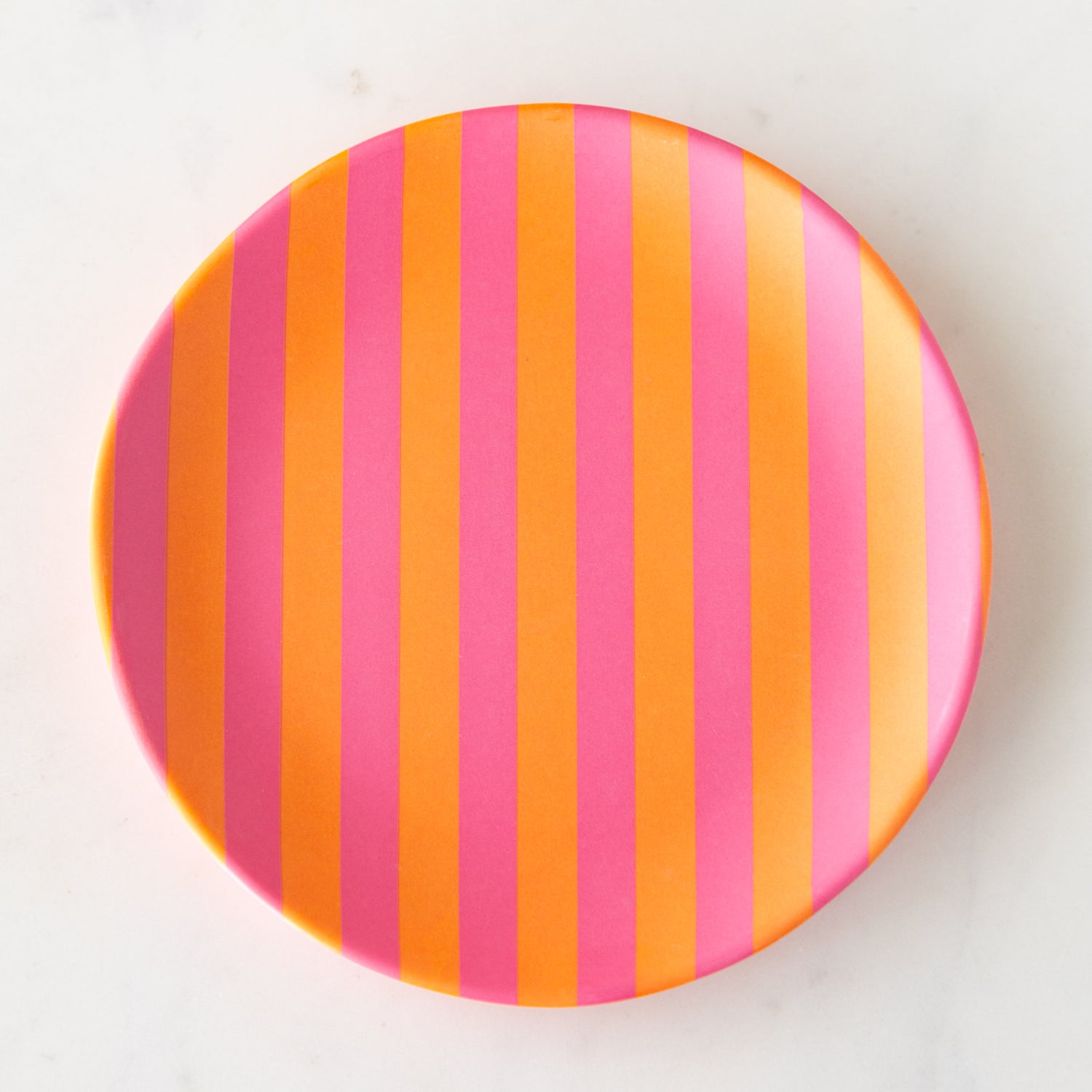 A round Stripe Melamine Plate with alternating vertical pink and orange stripes on a marble table.