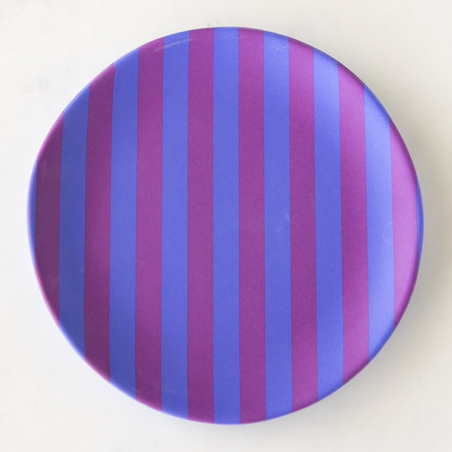 A round Stripe Melamine Plate with alternating vertical purple and blue stripes on a marble table.