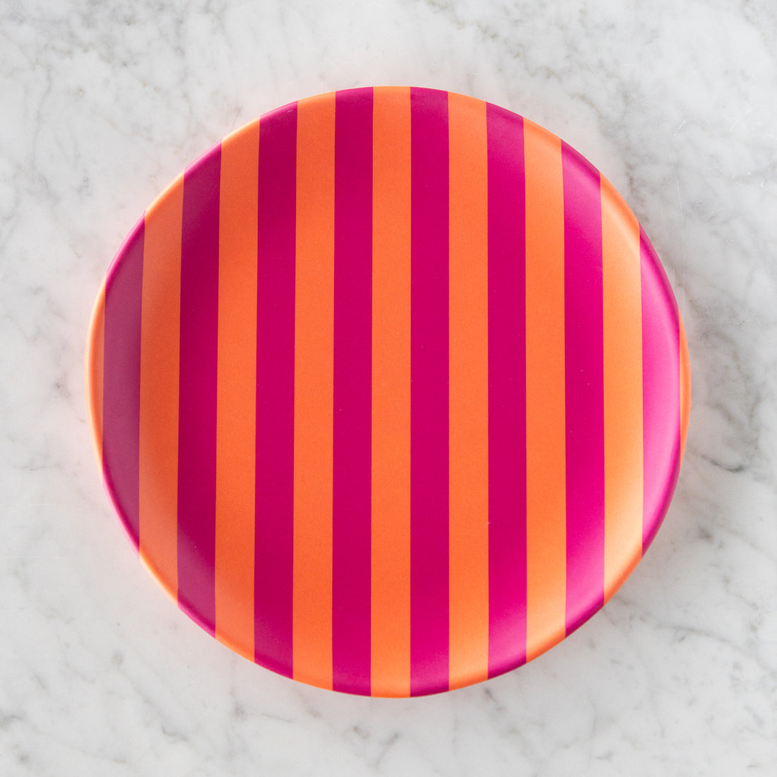A round Stripe Melamine Plate with alternating vertical orange and magenta stripes on a marble table.