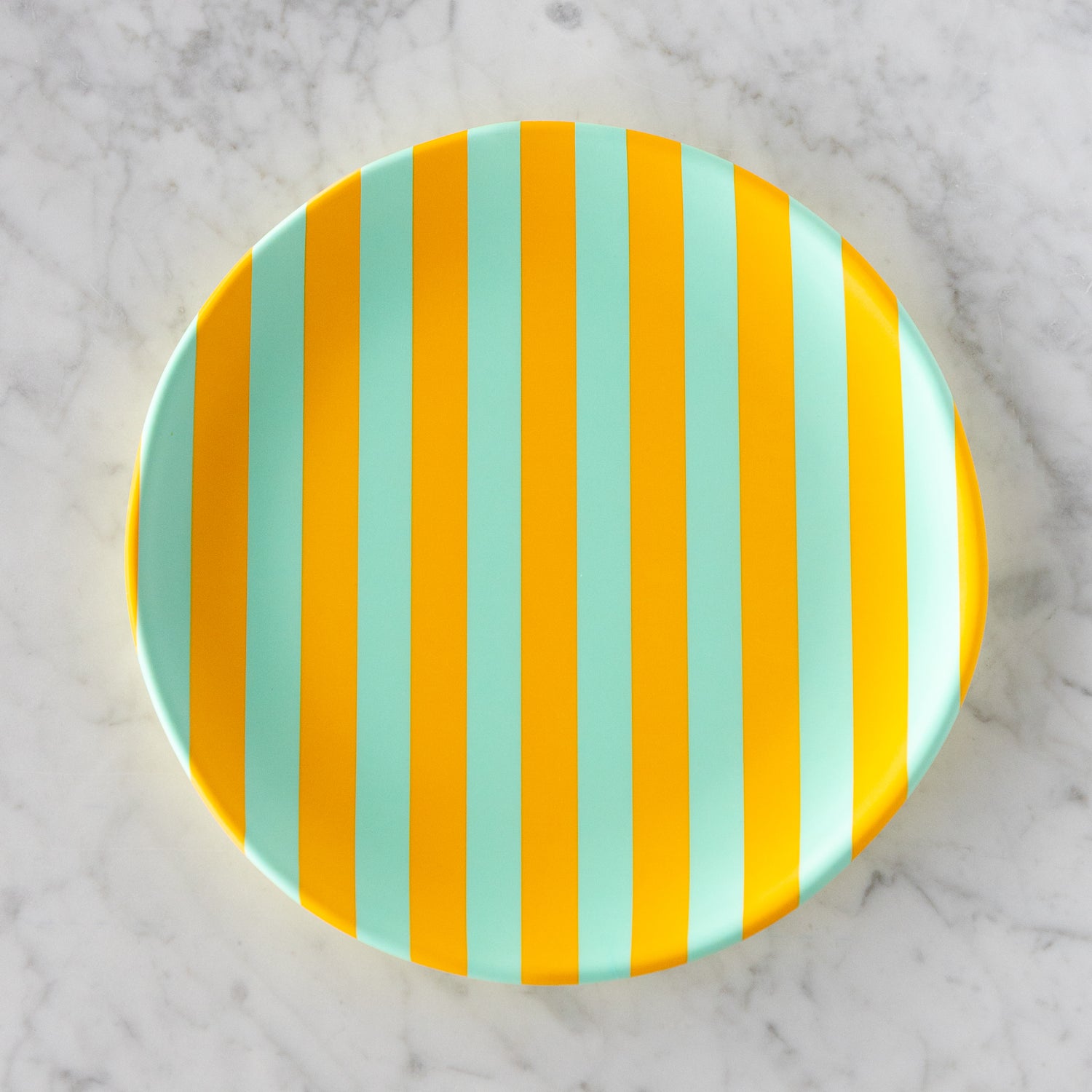 A round Stripe Melamine Plate with alternating vertical marigold and seafoam stripes on a marble table.