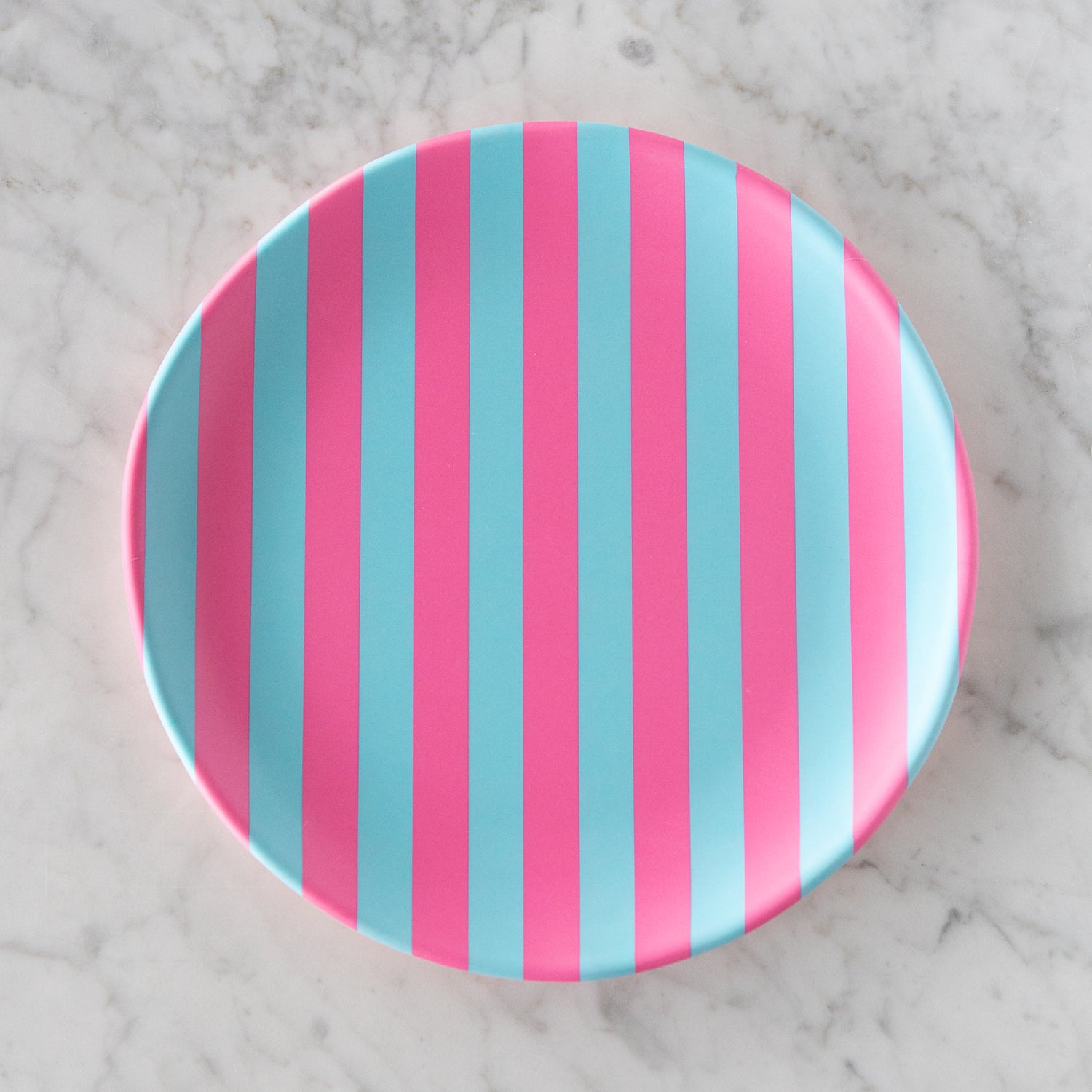 A round Stripe Melamine Plate with alternating vertical pink and light blue stripes on a marble table.