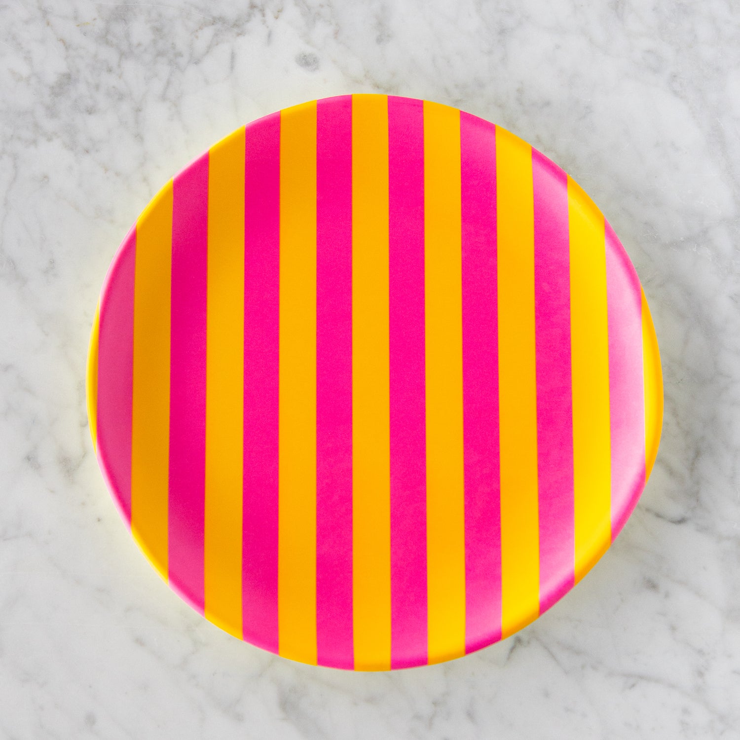 A round Stripe Melamine Plate with alternating vertical hot pink and yellow stripes on a marble table.