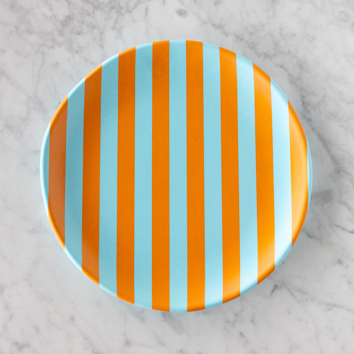 A round Stripe Melamine Plate with alternating vertical light blue and orange stripes on a marble table.