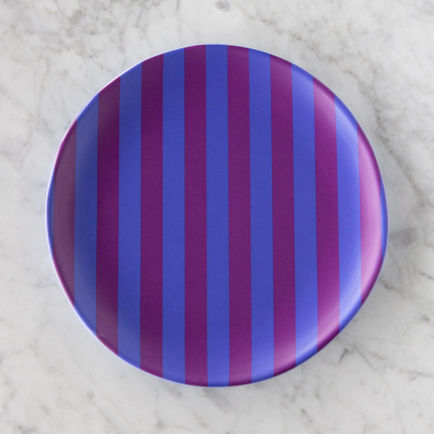 A round Stripe Melamine Plate with alternating vertical purple and blue stripes on a marble table.