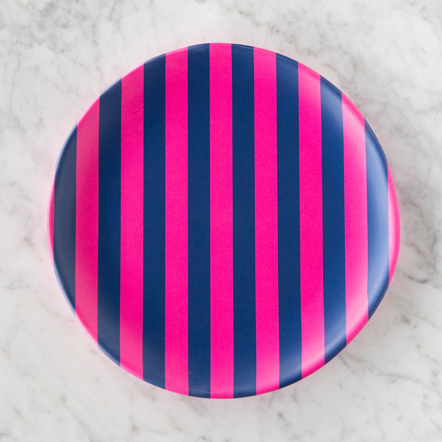 A round Stripe Melamine Plate with alternating vertical hot pink and navy stripes on a marble table.