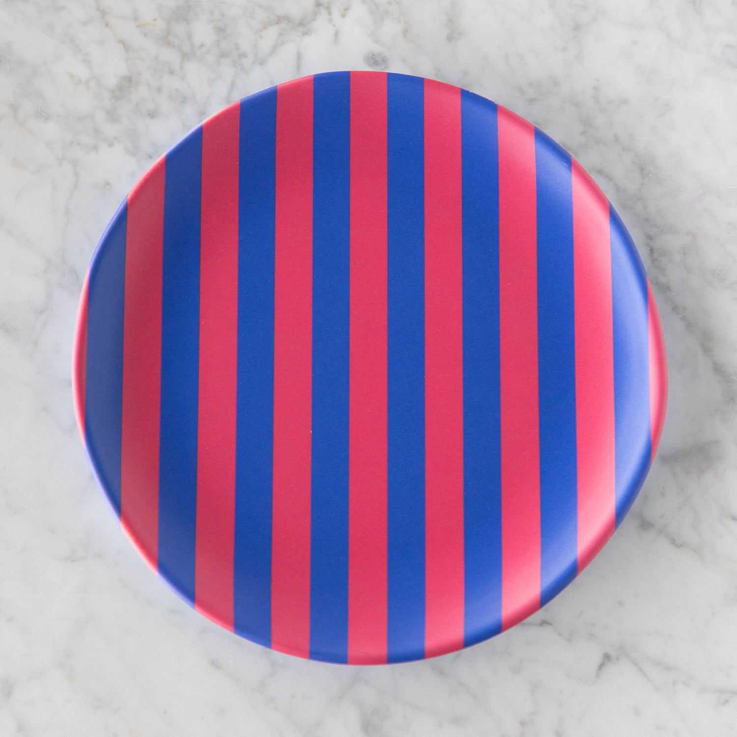 A round Stripe Melamine Plate with alternating vertical pink and blue stripes on a marble table.