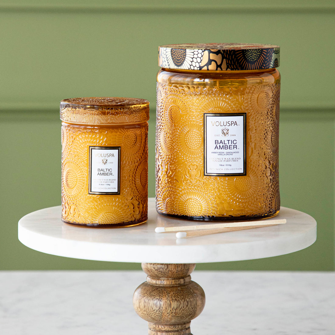 Two Voluspa Baltic Amber candles with matches on a marble surface.