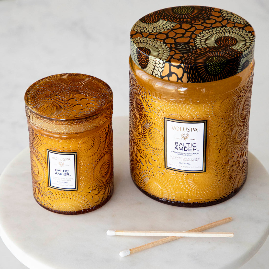 Two Voluspa Baltic Amber candles with matches on a marble surface.