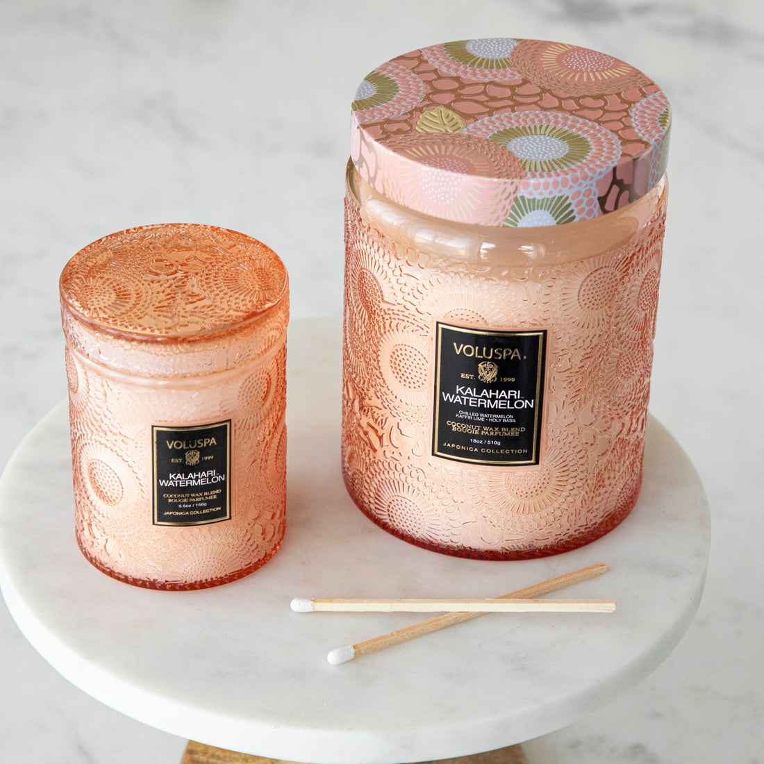 Two Voluspa Kalahari Watermelon candles on a marble surface with matches nearby, featuring a crisp cucumber aroma.